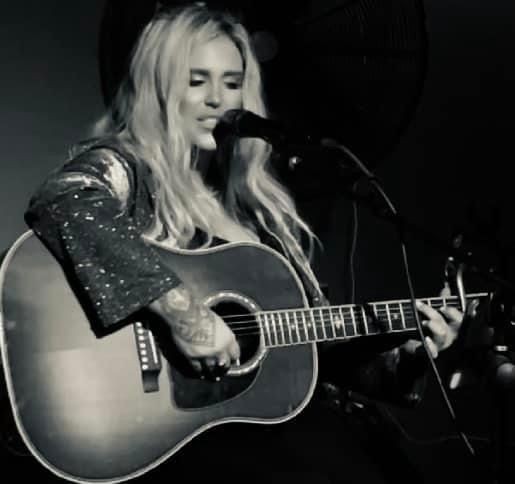 Person with long hair and tattoos playing an acoustic guitar on stage under dark, dramatic lighting.